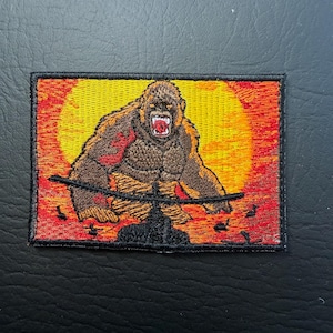 May include: Embroidered patch depicting a brown gorilla with an open mouth, set against a yellow and orange sunset. The gorilla is positioned above a black line, with a black border. The patch is rectangular.