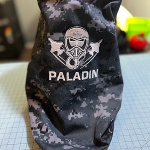 May include: A black and grey camouflage fabric bag with a white embroidered design of a pilot wearing a helmet and goggles with two fighter jets on either side. The text "PALADIN" is embroidered below the design.