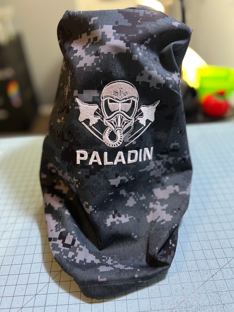 May include: A black and grey camouflage fabric bag with a white embroidered design of a pilot wearing a helmet and goggles with two fighter jets on either side. The text "PALADIN" is embroidered below the design.