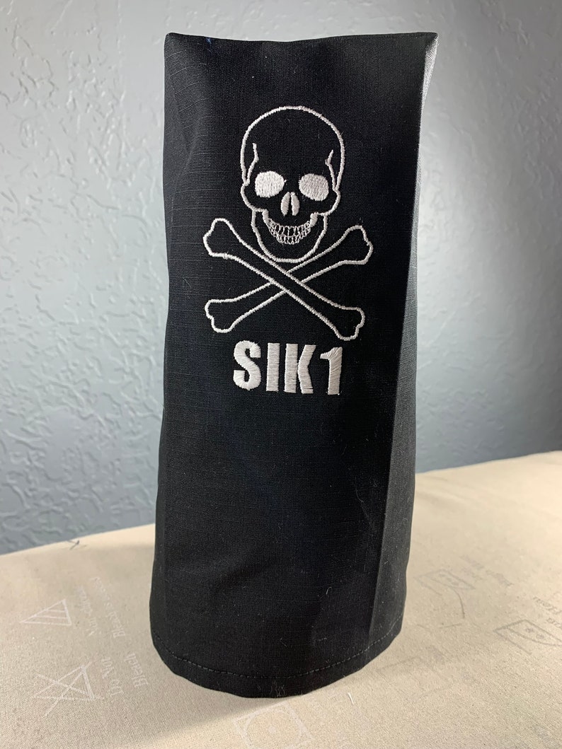 May include: Black golf club head cover featuring a white embroidered skull and crossbones design. The text "SIK1" is embroidered below the skull and crossbones.