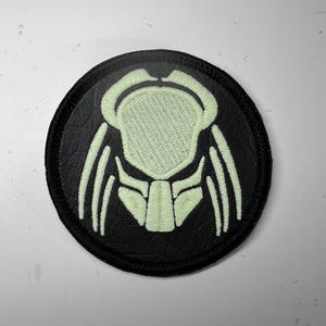 May include: A circular black patch with a light green embroidered Predator mask design. The patch has a black border and a textured appearance. The Predator mask design is detailed and stylized.