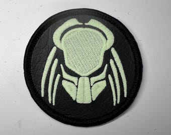 Op de afbeelding: Een ronde, zwarte patch met een lichtgroen geborduurd ontwerp van een Predator-masker. De patch heeft een zwarte rand en een getextureerd uiterlijk. Het Predator-maskerontwerp is gedetailleerd en gestileerd.