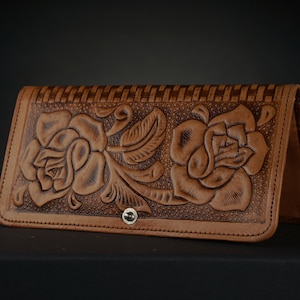 Puede incluir: Una cartera de cuero marrón con un diseño floral en relieve en la parte delantera. El diseño presenta dos rosas con detalles intrincados y hojas.