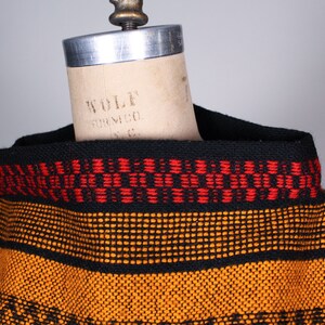 May include: A black and orange striped sweater with a red and black patterned trim around the neck. The sweater is on a dress form.