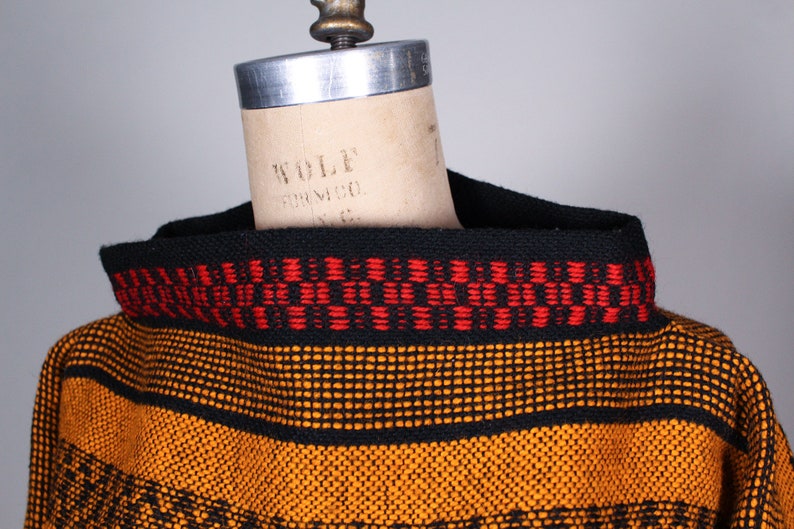 May include: A black and orange striped sweater with a red and black patterned trim around the neck. The sweater is on a dress form.