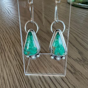 May include: A pair of silver earrings with a triangular, vibrant green stone. The stones are set in a silver frame with a decorative silver design at the bottom. The earrings hang from a silver hoop and hook.