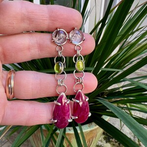 May include: A pair of dangle earrings with a teardrop-shaped ruby gemstone, a green gemstone, and a clear gemstone with black inclusions. The earrings are made of silver.