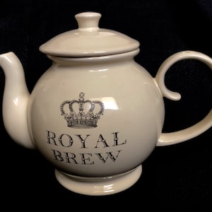 Könnte beinhalten: Eine cremefarbene Teekanne aus Keramik mit einer schwarzen Krone und dem Aufdruck "Royal Brew" auf der Seite.