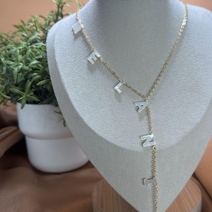 May include: A gold chain necklace with the name "ELANIE" spelled out in white letters. The necklace is displayed on a white mannequin bust with a wooden base.