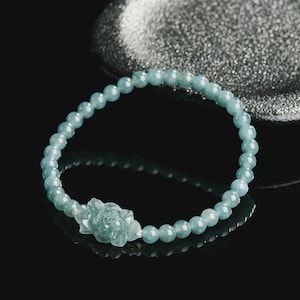 Puede incluir: Una pulsera de cuentas azul claro con una flor tallada en el centro. La pulsera presenta pequeñas cuentas redondas y un diseño floral detallado. La joya se exhibe sobre una superficie negra reflectante, con un fondo plateado borroso.