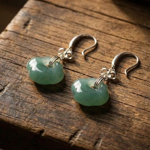 Puede incluir: Un par de pendientes de jade con ganchos de plata. Los pendientes presentan una piedra de jade verde claro en forma de rosquilla. Se exhiben sobre una superficie de madera rústica.