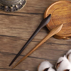 May include: Two wooden hair sticks. One is dark brown, the other is light brown. Both are long and slender with pointed ends. They are displayed on a wooden surface with a decorative plate and cotton.