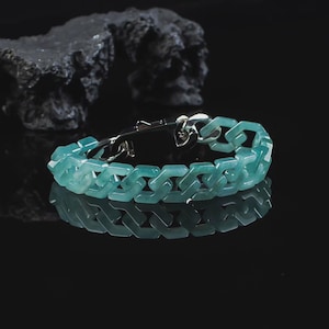 Puede incluir: Una pulsera turquesa con eslabones de cadena y un cierre plateado. La pulsera está hecha de eslabones rectangulares translúcidos. La pulsera se muestra sobre una superficie negra reflectante.