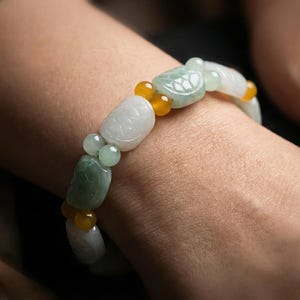 Puede incluir: Un brazalete de jade con cuentas verdes, blancas y amarillas. El brazalete presenta varias formas, incluyendo cuentas redondas y oblongas, con un diseño floral tallado en una de las cuentas más grandes. El brazalete se lleva en la muñeca.