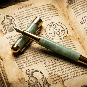May include: A vintage-style fountain pen with a green marbled barrel and gold accents rests on an open book. The book's pages are aged, with handwritten text and illustrations, including the word "ALCHEMY".