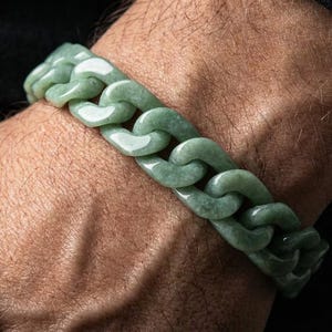 Puede incluir: Una pulsera de cadena de jade verde claro. La pulsera está hecha de eslabones entrelazados y se usa en la muñeca. El fondo es una tela oscura.