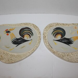 May include: Two ceramic wall plaques in the shape of a rooster with a textured, off-white background. The roosters are painted in black, yellow, green, and red.