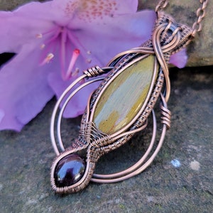 May include: A copper wire wrapped pendant featuring a green and brown stone with a smaller garnet stone at the bottom.