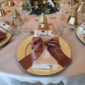 May include: A gold charger plate with a brown velvet ribbon tied in a bow with the name "Sophie" written on it. The plate is set on a white tablecloth with gold and white decorations.