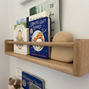 May include: A wooden shelf with a light brown finish, mounted on a white wall. The shelf has a metal bar across the front and is holding a stack of children's books. The books have colorful covers with illustrations of animals and text. The top book is titled "Giraffes Can't Dance".