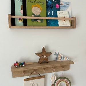 May include: A wooden shelf with a star-shaped wooden sign that says "Aila" and a pennant that says "Happy". The shelf has hooks on the bottom and is decorated with a rainbow, a hot air balloon, and a sign that says "Sleep little".