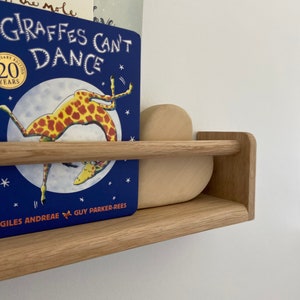 May include: A wooden shelf with a blue book titled "Giraffes Can't Dance" by Giles Andreae and Guy Parker-Rees. The book is on the top shelf and a wooden heart-shaped object is on the bottom shelf.