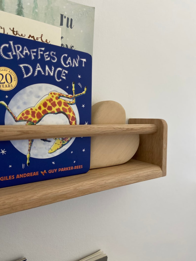 May include: A wooden shelf with a blue book titled "Giraffes Can't Dance" by Giles Andreae and Guy Parker-Rees. The book is on the top shelf and a wooden heart-shaped object is on the bottom shelf.