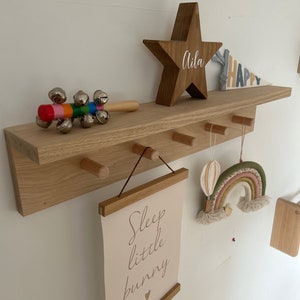 May include: A wooden shelf with a star-shaped wooden sign that says "Aila" and a rainbow-colored felt rainbow hanging from a hook. There is also a sign that says "Sleep little bunny" and a wooden toy with bells.