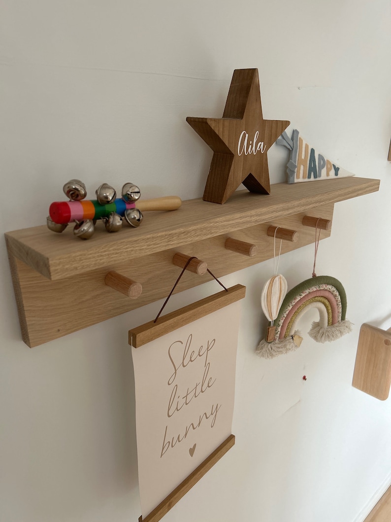 May include: A wooden shelf with a star-shaped wooden sign that says "Aila" and a rainbow-colored felt rainbow hanging from a hook. There is also a sign that says "Sleep little bunny" and a wooden toy with bells.