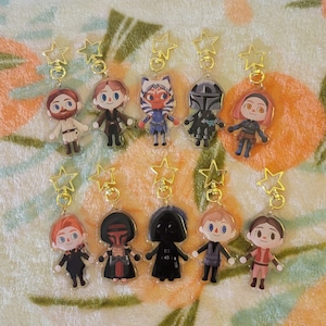 Puede incluir: Colección de llaveros de personajes con cierres dorados en forma de estrella. Los llaveros presentan varios personajes en un estilo de dibujos animados. Los personajes visten atuendos con colores como negro, azul, rojo y blanco. El fondo es una tela estampada.