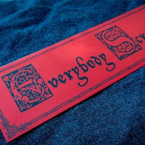 Könnte beinhalten: Ein rechteckiges rotes Etikett mit den Worten "everybody cream!" in dunkelblauer Schrift. Das Etikett hat dekorative Eckendesigns und zwei verzierte Buchstabendesigns. Das Etikett ist vor einem dunkelblauen strukturierten Hintergrund platziert.