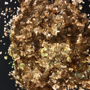 May include: A pile of iridescent brown and gold mica flakes. The flakes are small and irregular in shape, and they have a shimmery, pearlescent finish.