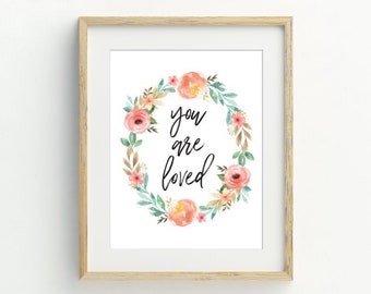 Könnte beinhalten: Ein gerahmter Druck mit einem Aquarell-Blumenkranz in Pfirsich-, Rosa- und Grüntönen. Die Worte "you are loved" sind in eleganter Schrift innerhalb des Kranzes geschrieben. Der Rahmen hat eine helle Holzfarbe.