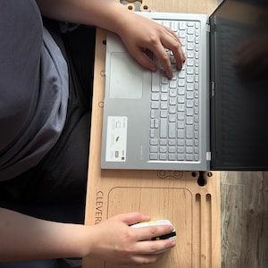 May include: A wooden laptop stand with a built-in mouse pad and a groove for a phone. The stand has the word "CLEVERL" engraved on it. A person is using a laptop and a wireless mouse on the stand.