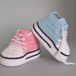 Puede incluir: Un par de zapatillas de bebé hechas a mano de estilo botín. Una es rosa claro, la otra azul claro. Ambas tienen suelas blancas, cordones blancos y rayas negras. Los zapatos están hechos de ganchillo.