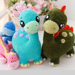May include: A group of handmade crochet dinosaur toys in different colours. The dinosaurs include a blue one with purple spots, a green one with red accents, a pink one with a yellow spine, and a blue one. Each toy has large, expressive eyes.