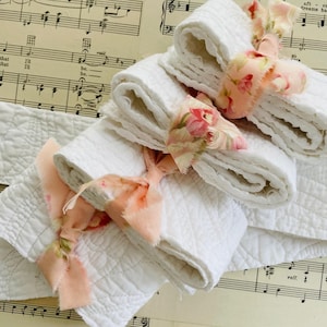 May include: White quilted fabric rolls tied with peach and floral patterned ribbons. The fabric is folded and stacked, creating a layered effect. The background features sheet music.