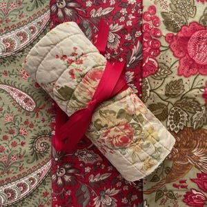 May include: A rolled quilted fabric piece featuring a floral pattern in cream, pink, and green, tied with a bright red ribbon. It rests on three distinct fabric panels: one with a green paisley print, one with a red floral design, and one with a large-scale floral and bird motif in green and pink.