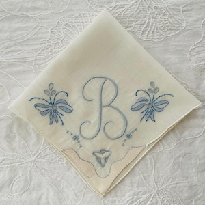 May include: A square off-white handkerchief featuring a central embroidered letter B in light blue thread. The design includes symmetrical light blue floral motifs on either side of the letter and a small decorative element at the bottom corner.