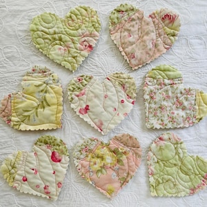 May include: Eight heart-shaped fabric ornaments featuring various floral and swirling quilted patterns in shades of soft green, pale pink, cream, and yellow. Each piece is stitched with a scalloped edge and detailed quilting lines across the surface.