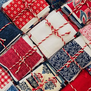 May include: A collection of stacked fabric squares tied with red and white baker's twine. The fabric bundles feature various patterns including red and white gingham, blue and white floral prints, solid red, and solid blue textures in a grid arrangement.