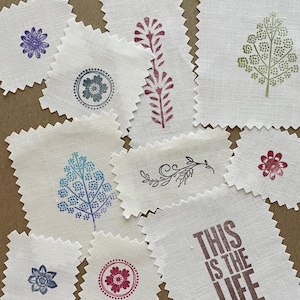 May include: A collection of fabric squares with various hand-stamped designs. The designs include flowers, leaves, and a simple swirl. The fabric is a natural linen color and has a slightly textured surface. One square has the text "This is the life" printed in brown.