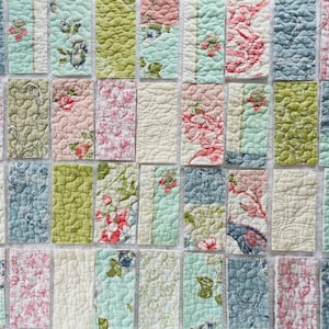 May include: A collection of rectangular fabric swatches featuring various quilted patterns in shades of sage green, dusty pink, cream, and muted blue. The fabric pieces display floral motifs, toile prints, and solid textures with visible stitching.