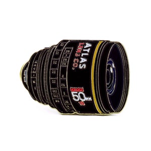 Puede incluir: Pin de metal dorado en forma de lente de cámara con el texto "ATLAS LENS CO. 50mm" en el lateral.