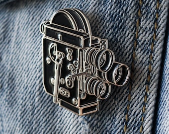 May include: Silver-toned metal pin shaped like a vintage movie camera. The camera is black with silver details and has multiple lenses. The pin is attached to a denim jacket.