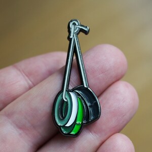 May include: A black metal pin with three stacked rings. The rings are green, white and black.