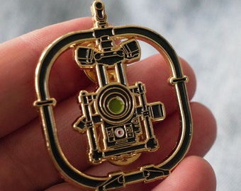 May include: Gold-toned metal pin featuring a black camera with a green lens, surrounded by a circular frame. The camera is detailed with intricate designs and appears to be a vintage style.