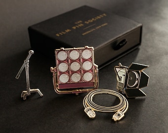 May include: Three gold-tone enamel pins depicting film set equipment. The pins feature a red and white film light, a black and white film light, and a black and white coiled cable.