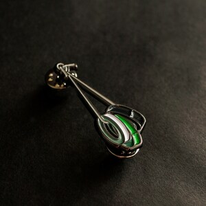May include: A silver pin featuring a green, white and black design. The pin is shaped like a pair of chopsticks with a round, green and white design at the end.