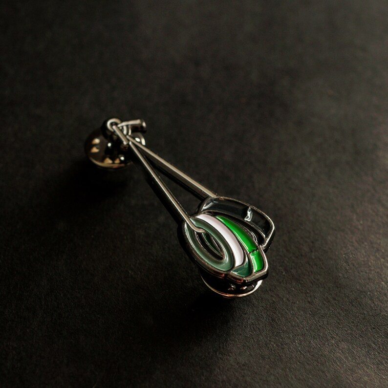 May include: A silver pin featuring a green, white and black design. The pin is shaped like a pair of chopsticks with a round, green and white design at the end.